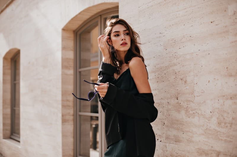 Home Cute Young Girl With Dark Wavy Hairstyle Bright Makeup Silk Dress Black Jacket Holding Sunglasses Hands Looking Away Against Beige Building Wall 1